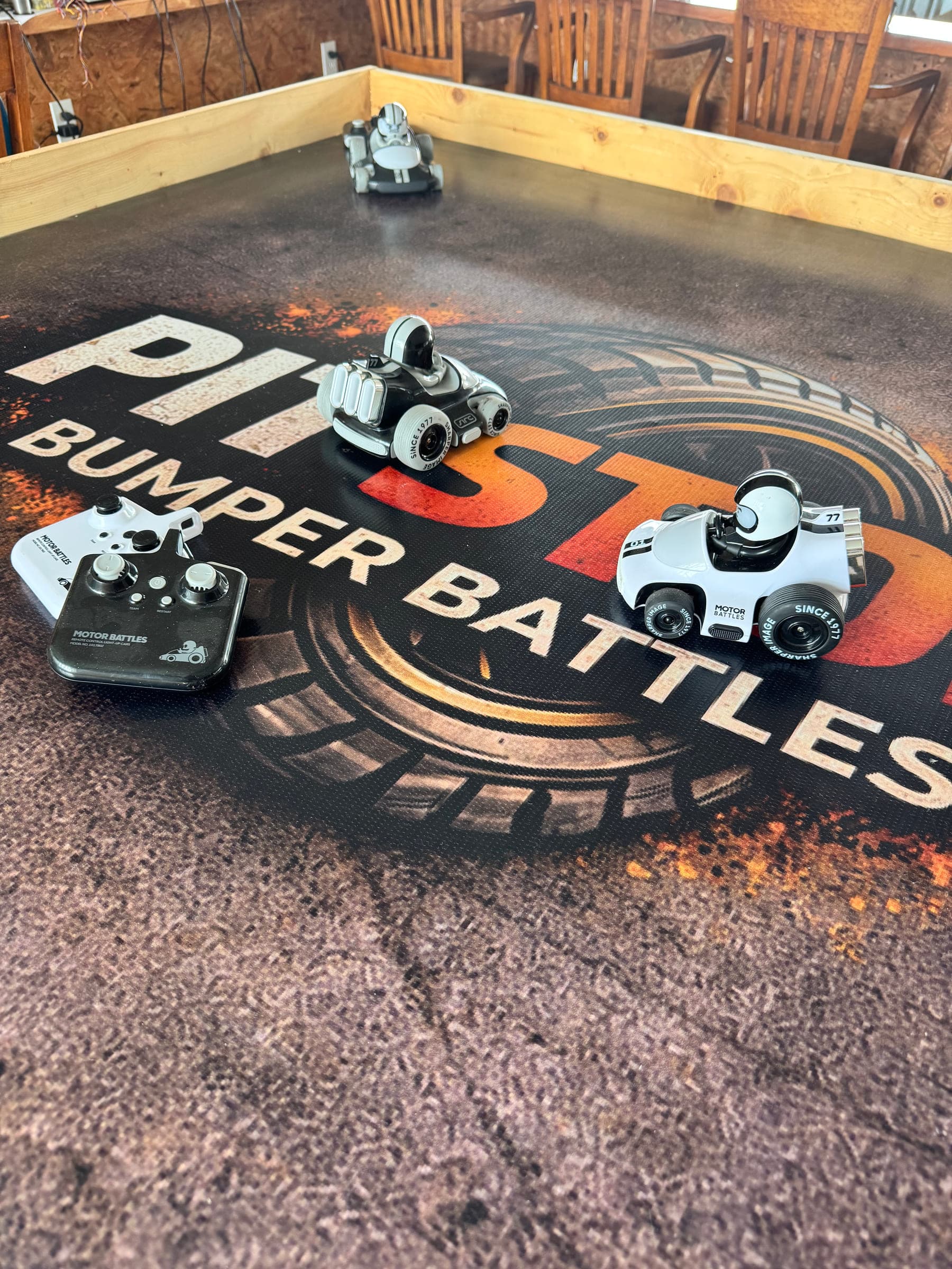 Bumper Battles en Pit Stop: dos carros de batalla a control remoto sobre el tapete de la arena con un control descansando a un lado.