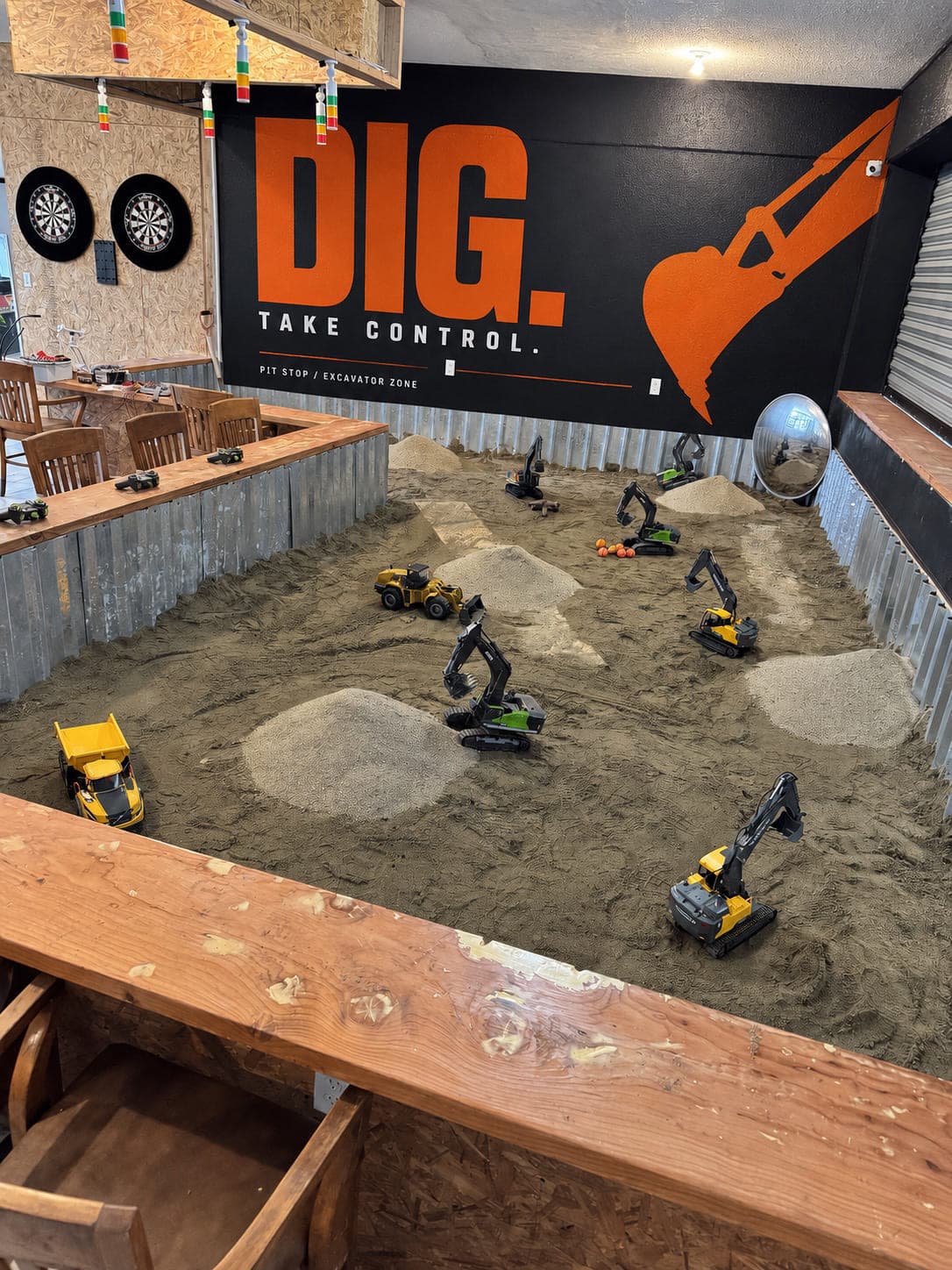 The Excavator Zone at Pit Stop: remote-control excavators and a loader working mounds of sand, with the DIG. TAKE CONTROL. mural on the wall behind and controllers waiting along the rail.