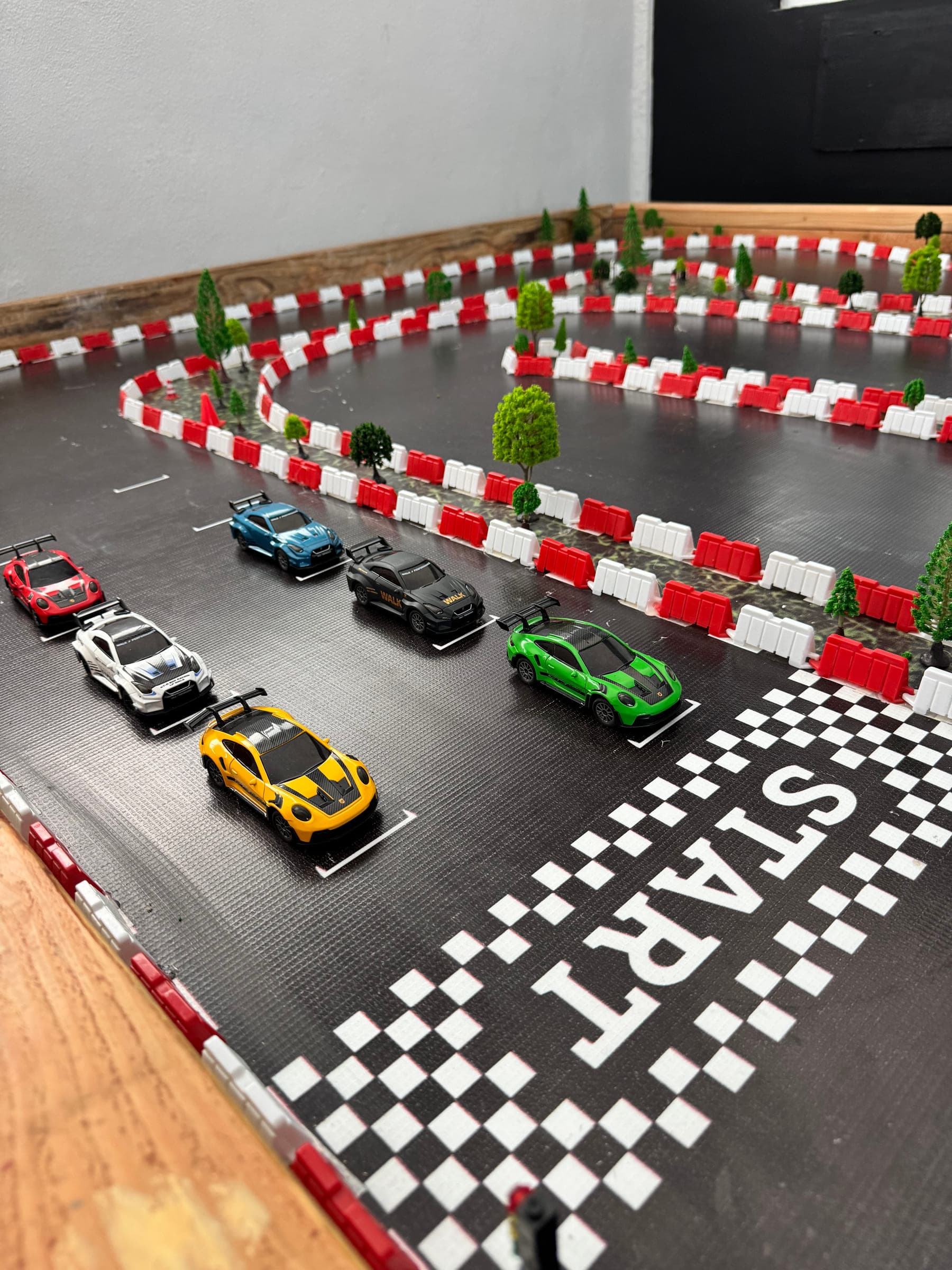 La pista RC de Pit Stop: carros formados en la parrilla detrás de la línea START pintada, con barreras rojas y blancas y árboles de maqueta alrededor del circuito.
