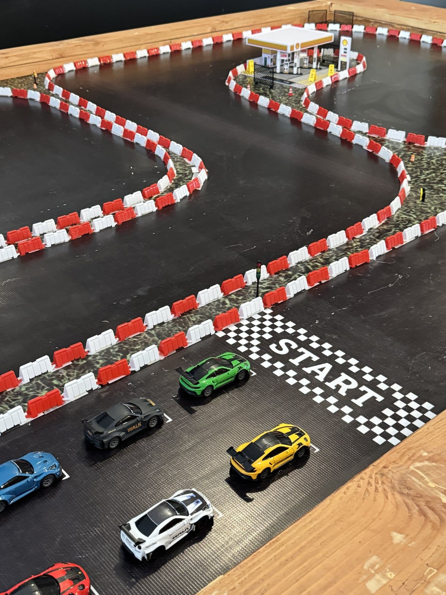 Vista cenital del circuito RC de Pit Stop: el trazado completo con sus dos curvas en S delimitadas por barreras rojas y blancas, la gasolinera de maqueta en la esquina y los carros formados detrás de la línea START.