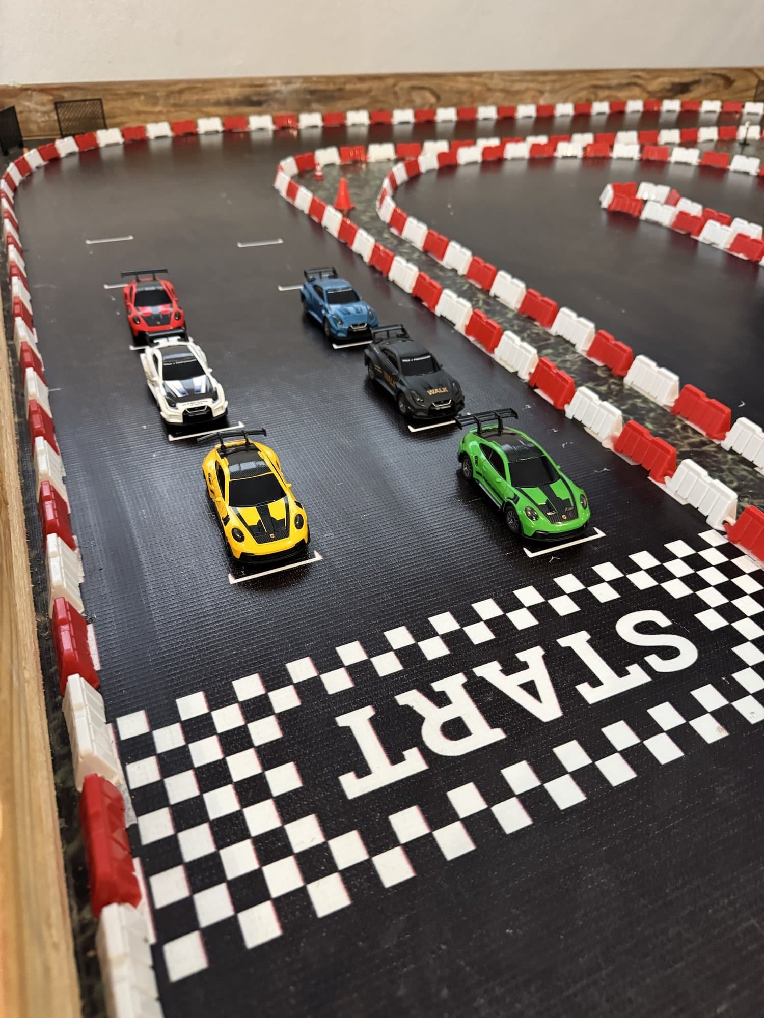 Carros formados en la parrilla de salida de la pista RC de Pit Stop, detrás de la línea START pintada, con barreras rojas y blancas alrededor del circuito.